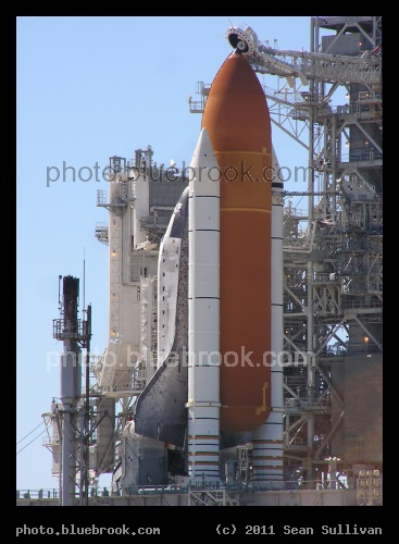 Behind the External Tank - Space shuttle Discovery at Kennedy Space Center launch pad 39-A, seen from a three-quarters angle northeast of the launch pad, showing the tiled underside of a wing, and the full height of the external fuel tank